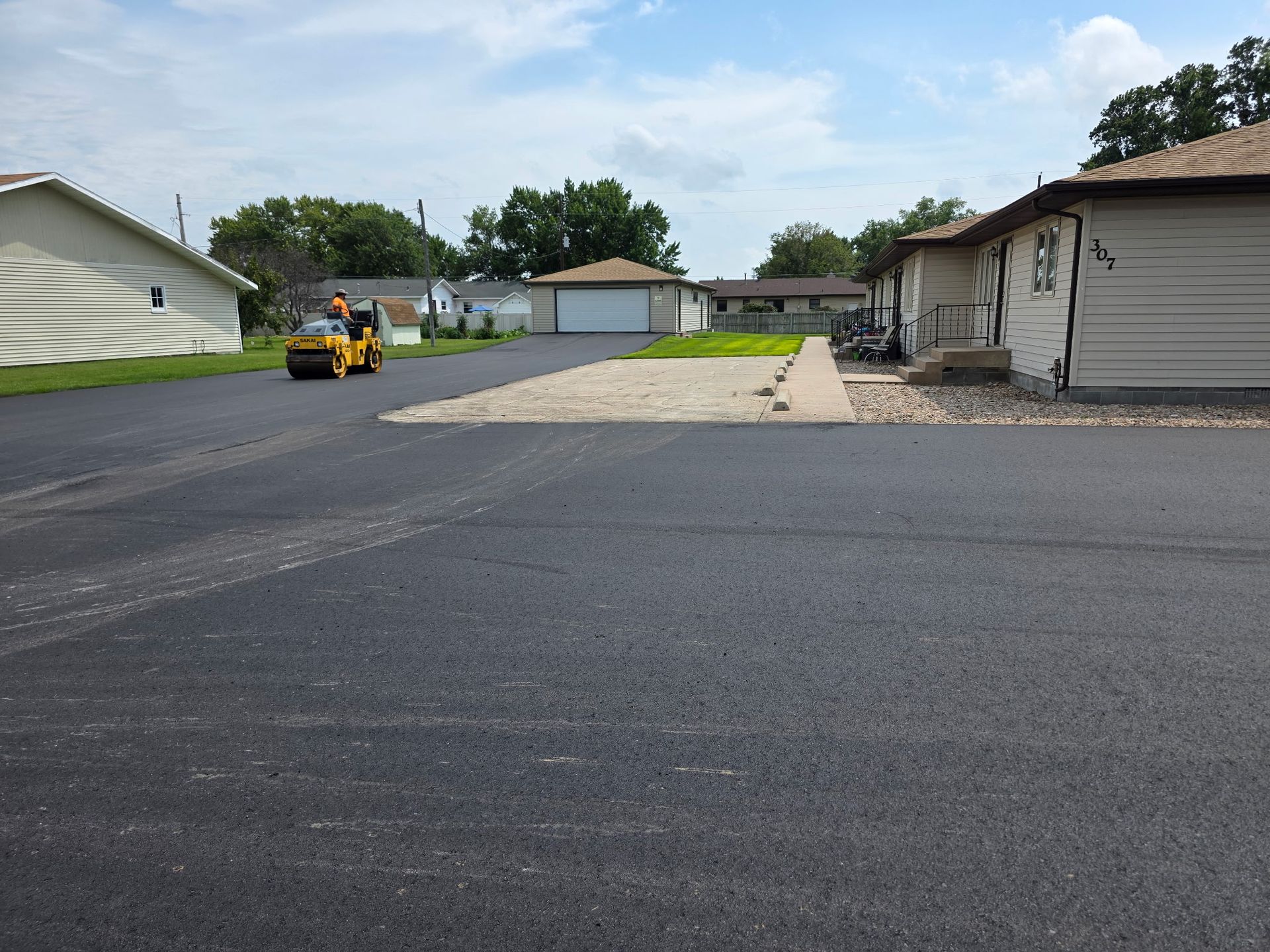 Asphalt paving work in progress: a small roller compacts fresh black asphalt near residential buildings.