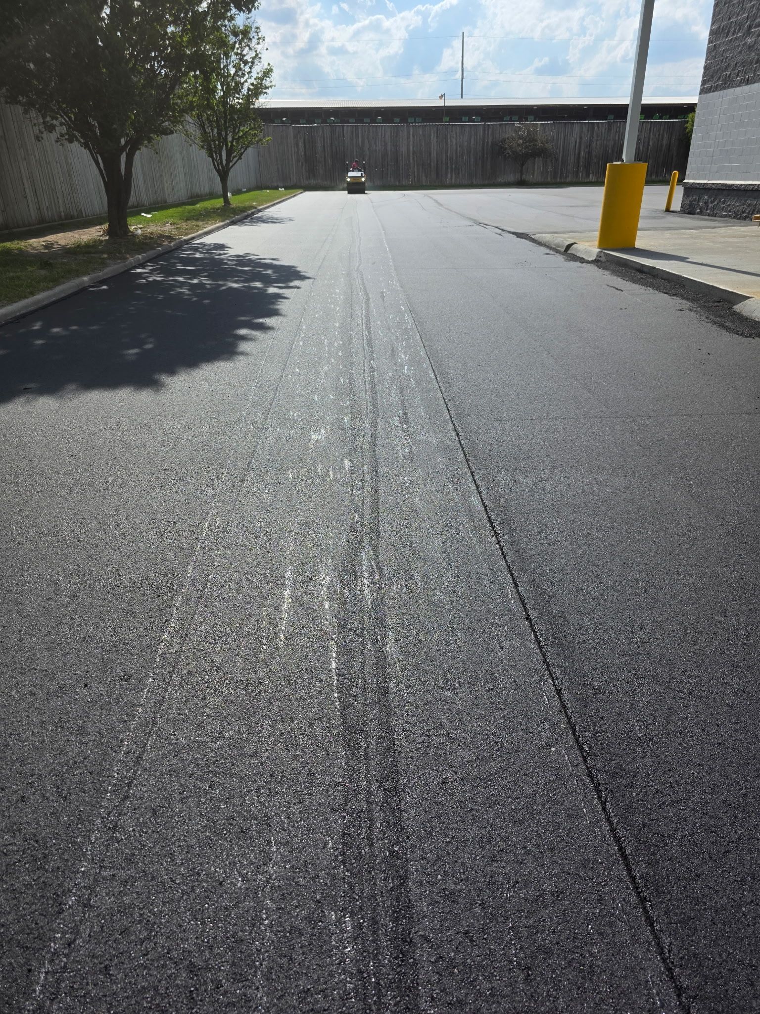 Newly paved asphalt road; sunlight reflects. Tree and fence in the background.