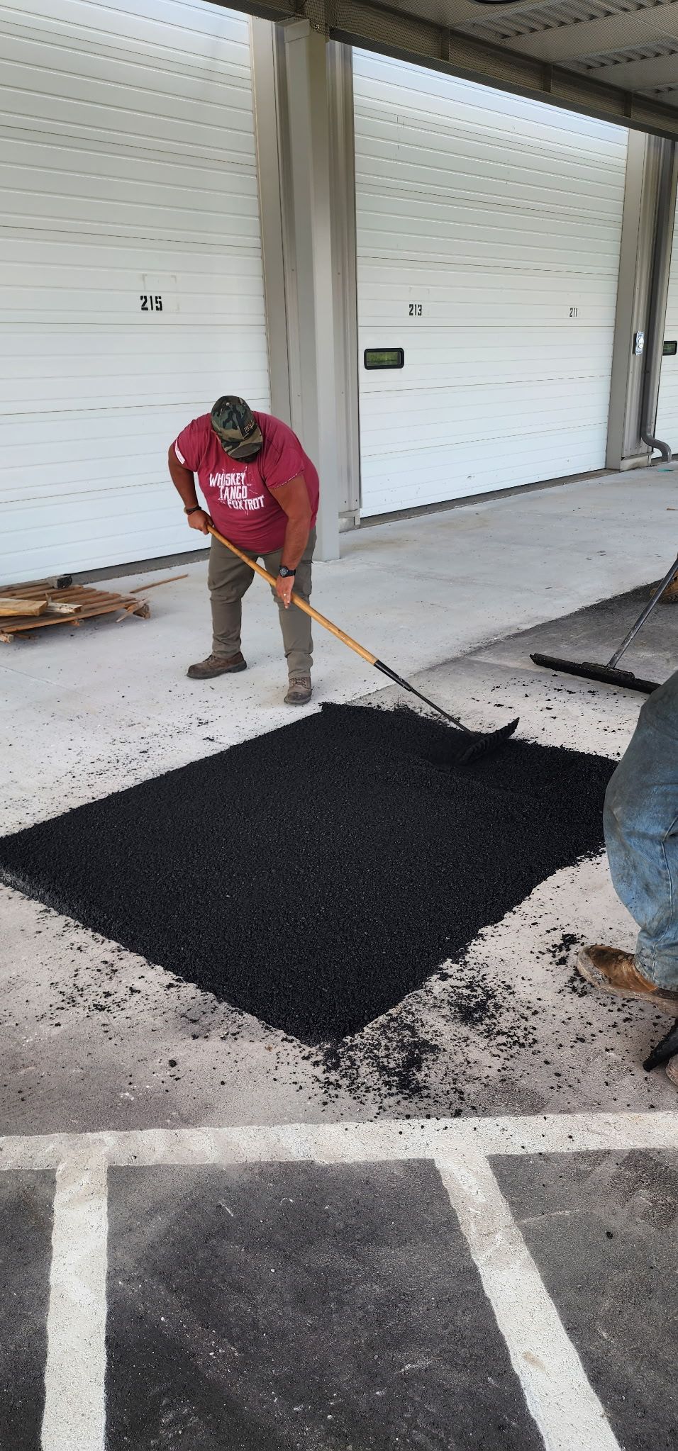 A person raking black asphalt on a concrete surface, likely repairing a parking lot.