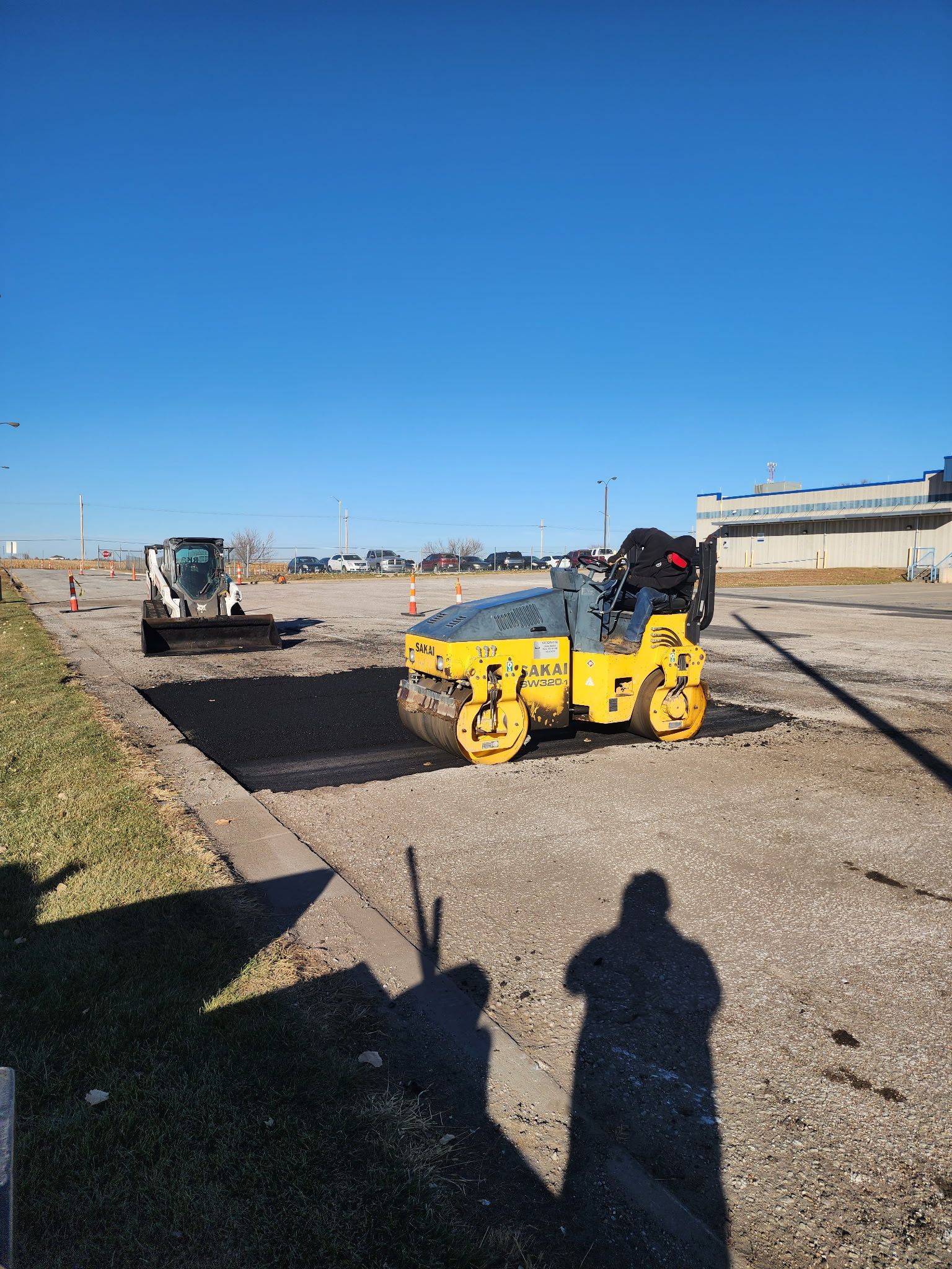 Road construction: Compactor rolling over gravel, Bobcat in background, clear sky.