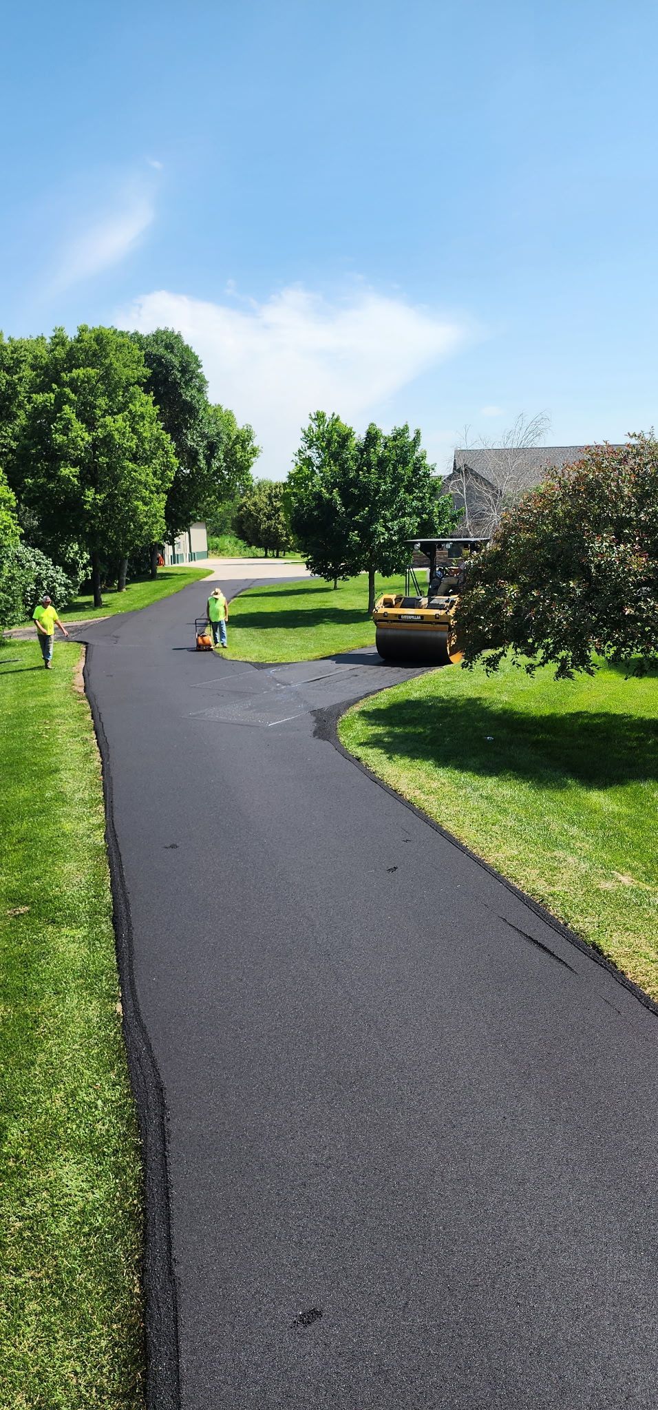 Asphalt path winding through grassy areas, trees under a blue sky.
