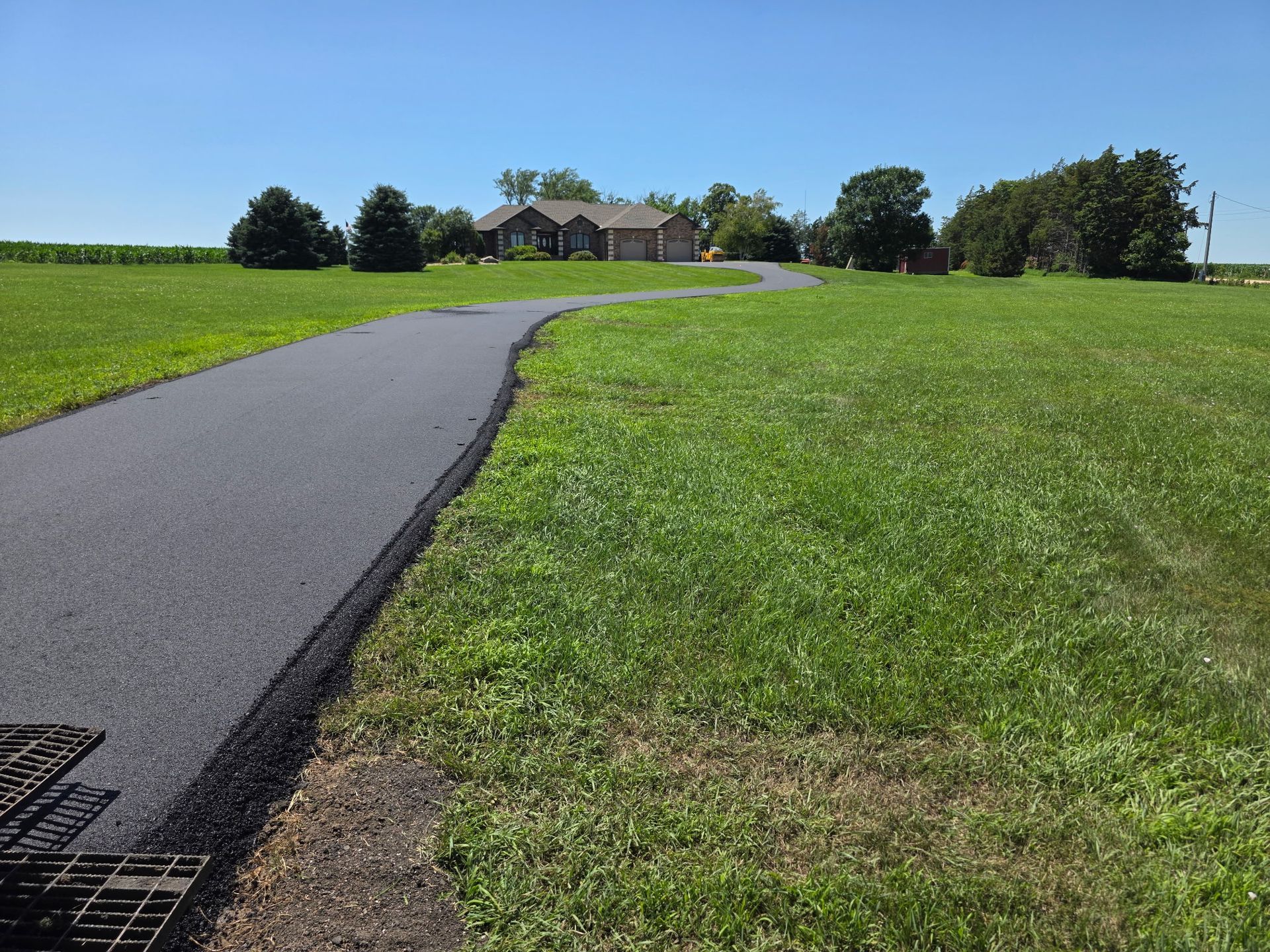 A paved driveway winds through a grassy field towards a house under a clear, blue sky.