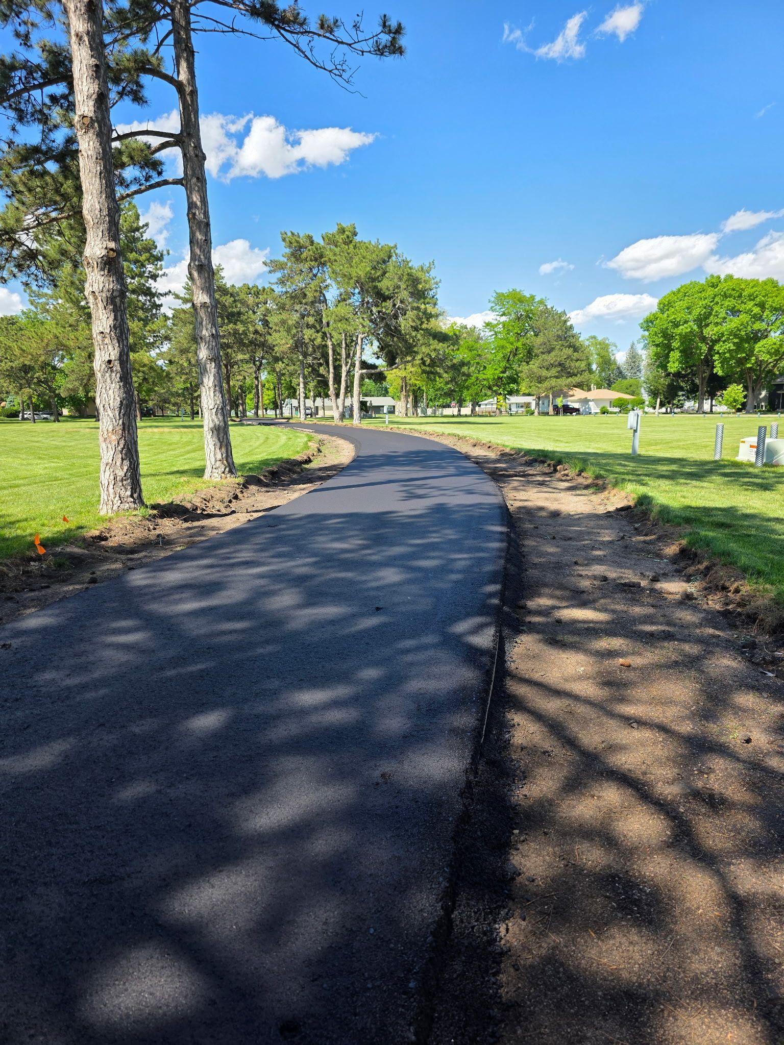 Newly paved asphalt pathway through a grassy park, trees on either side, blue sky.