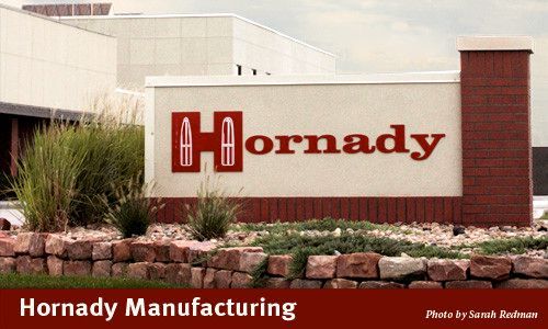 Sign for Hornady Manufacturing, red logo on white, brick base, building in background.