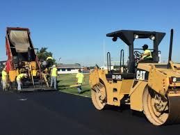 Construction worker in orange suit compacting asphalt with a yellow roller.