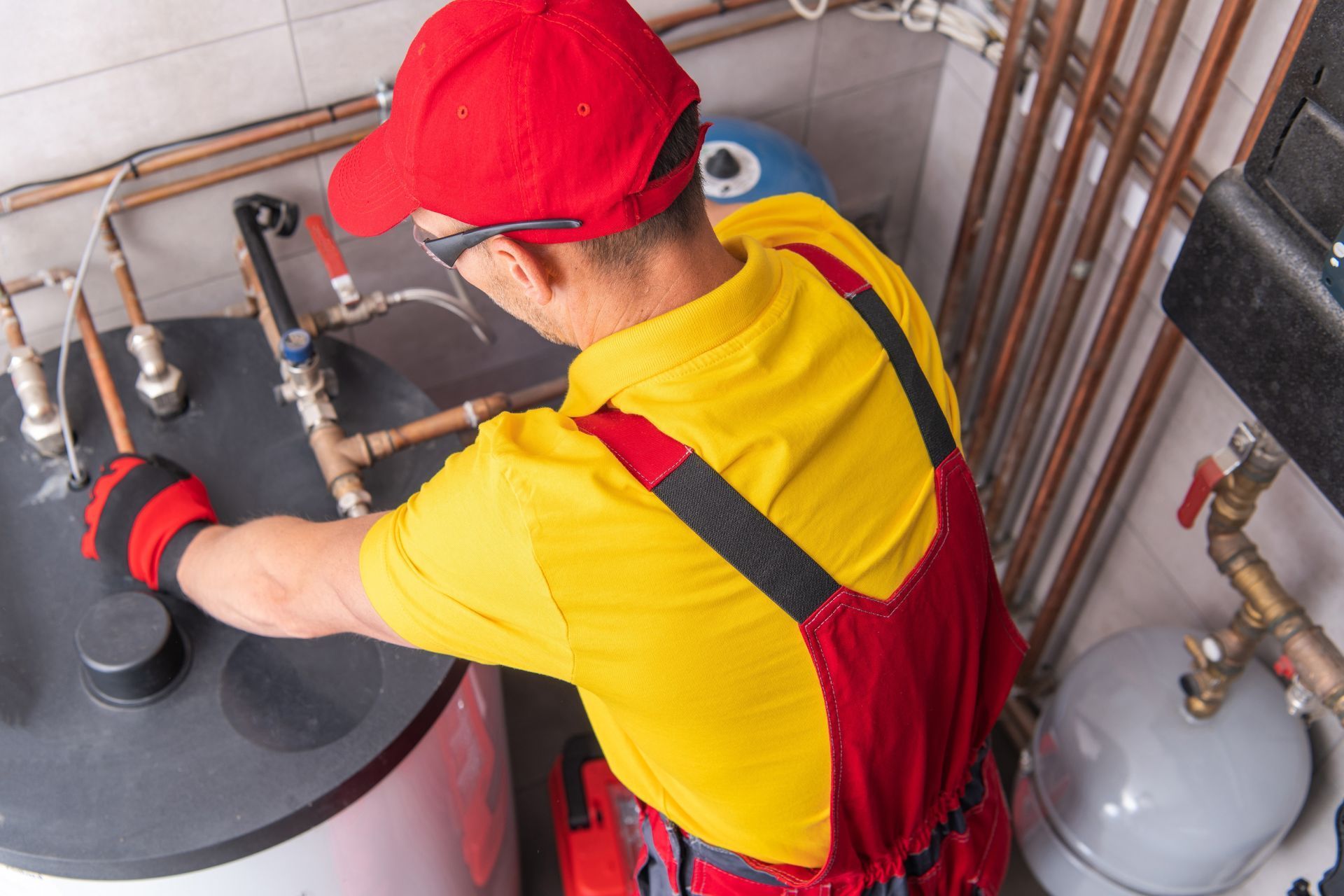 Plumber in red overalls and yellow shirt working on pipes in a utility room