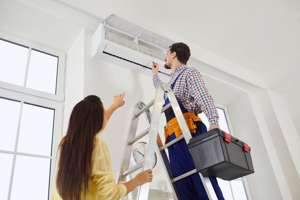 Two people installing a wall-mounted air conditioner, with one on a ladder holding tools.