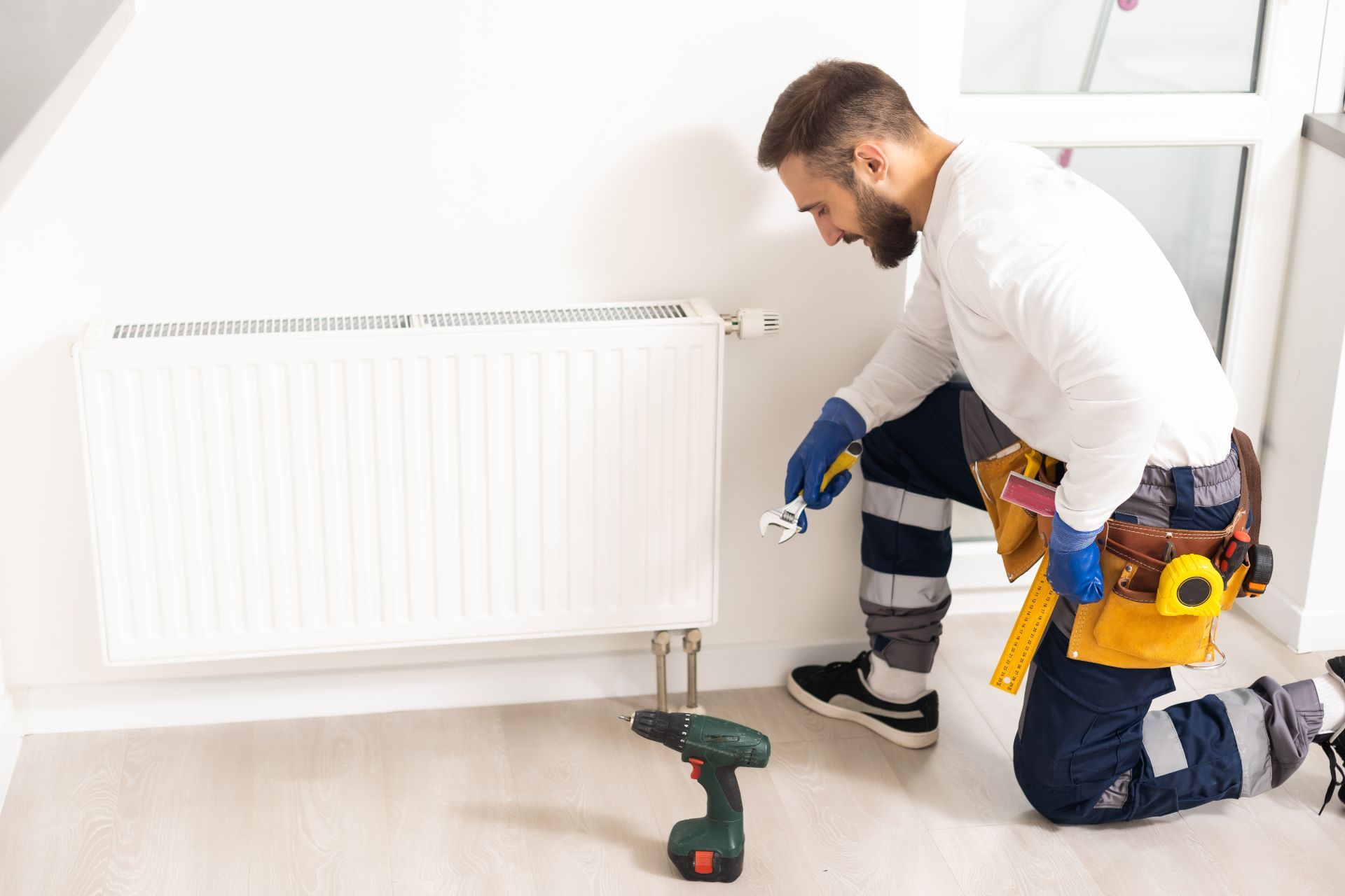 Plumber kneeling by a white radiator, tightening a valve with tools in a bright room.