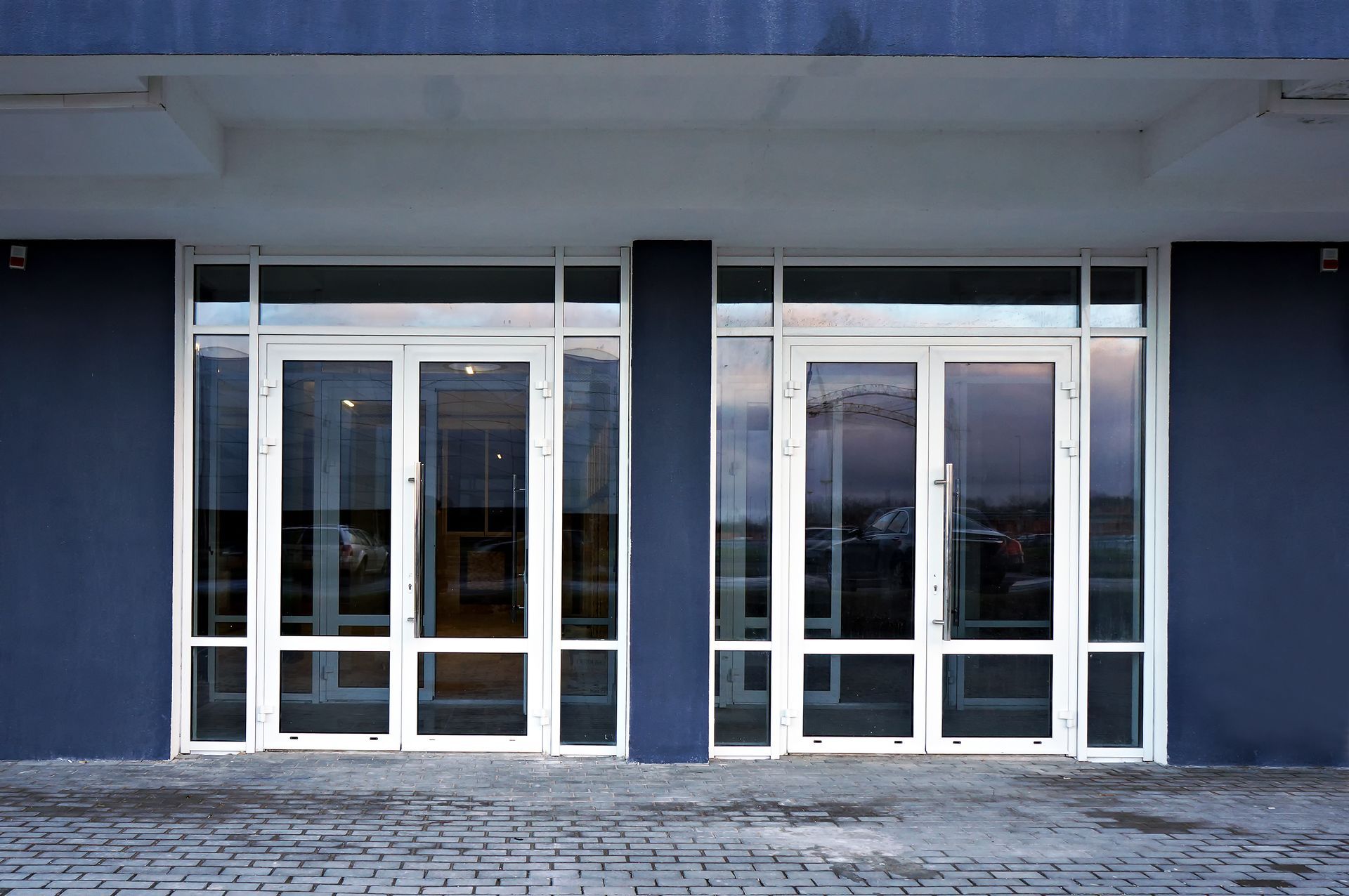 Double glass doors with white frames on a dark blue building with a gray brick ground.