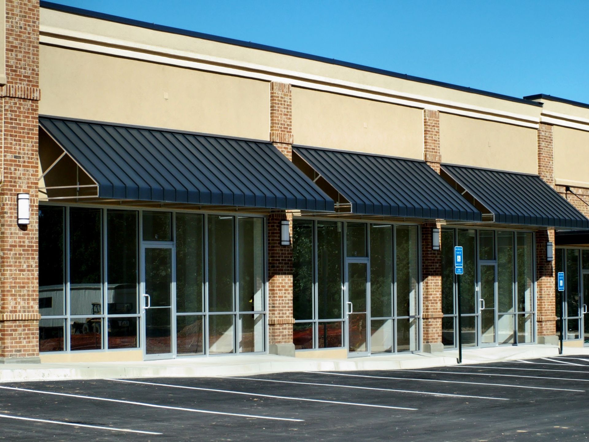 Empty storefronts with glass windows and black awnings, in a brick and tan building, on a sunny day.