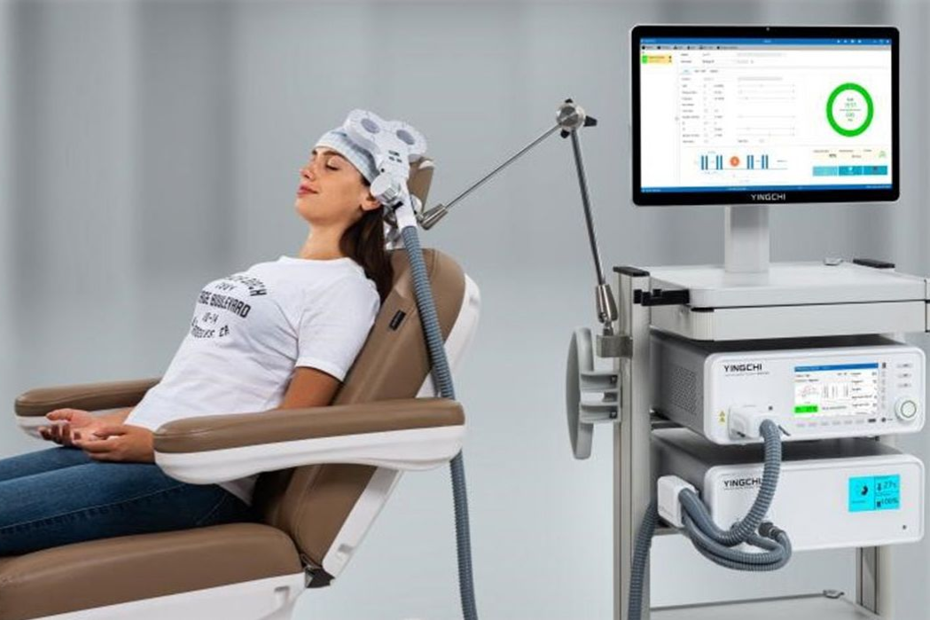 Woman in medical chair undergoing brain stimulation, with equipment monitoring on screen.