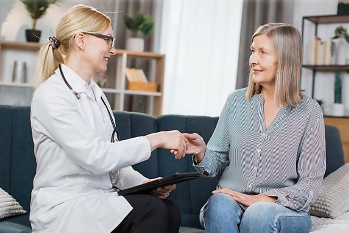 A doctor and patient fist bump on a sofa; inside a room with shelves and plants.