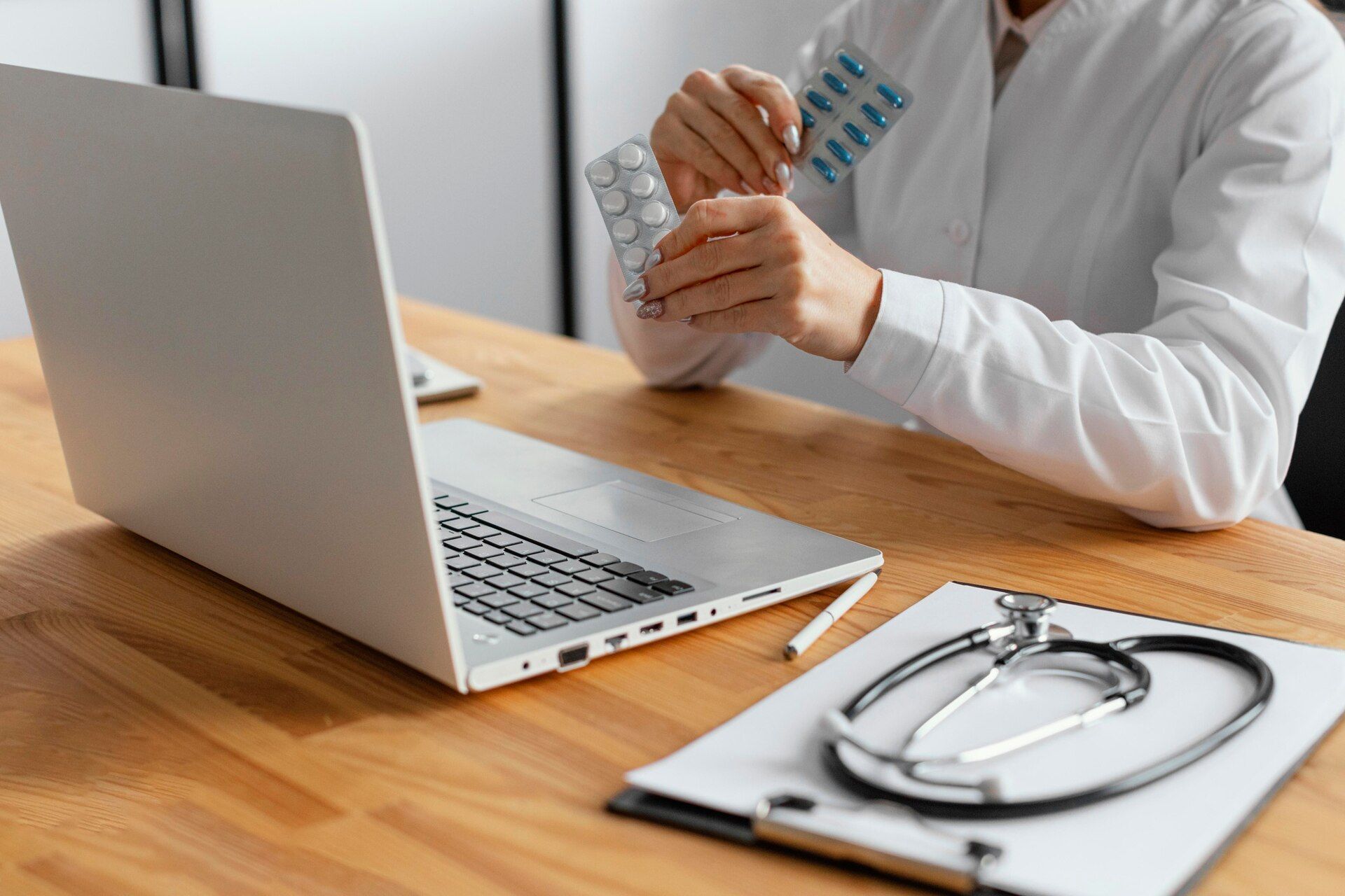 Doctor in a white coat holds pills while consulting on a laptop, with a stethoscope on a clipboard.