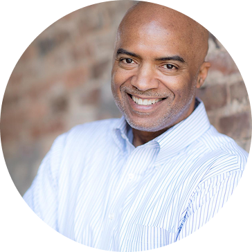Smiling man in a blue striped shirt against a brick wall.