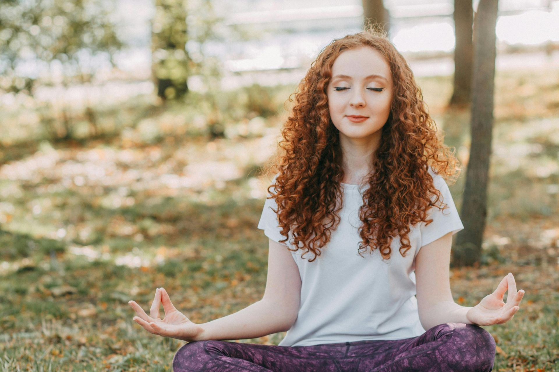 Woman with curly hair meditating outdoors, eyes closed, hands in yoga position, sunny park setting.