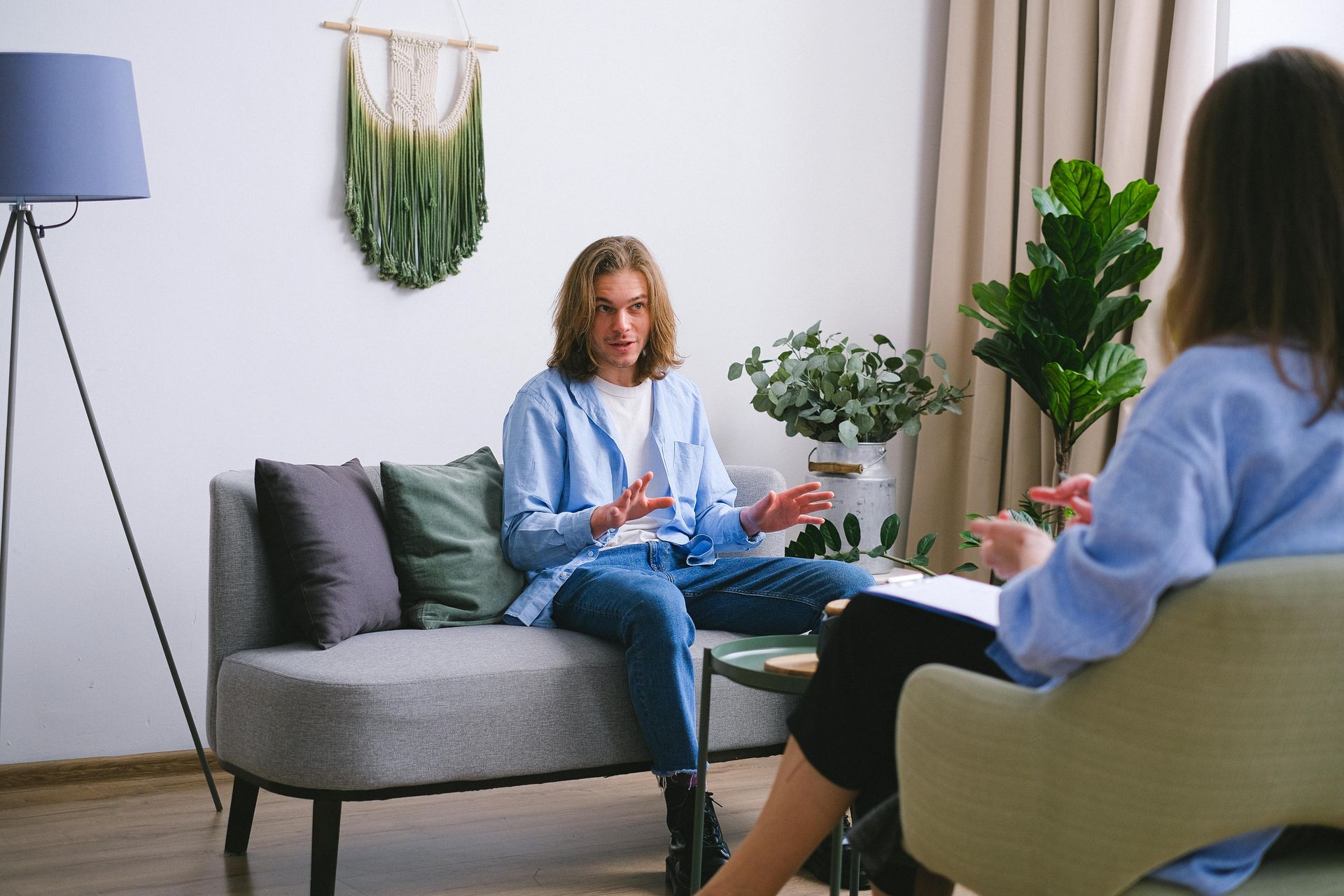A person seated on a couch gestures while talking to another person sitting in a chair, in an office.