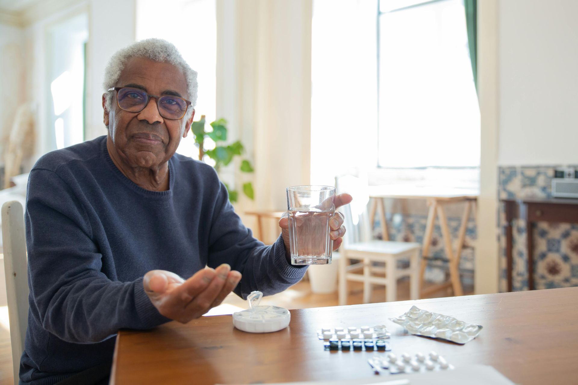 Older adult, wearing glasses, holds a glass of water, and pills sit on a table.