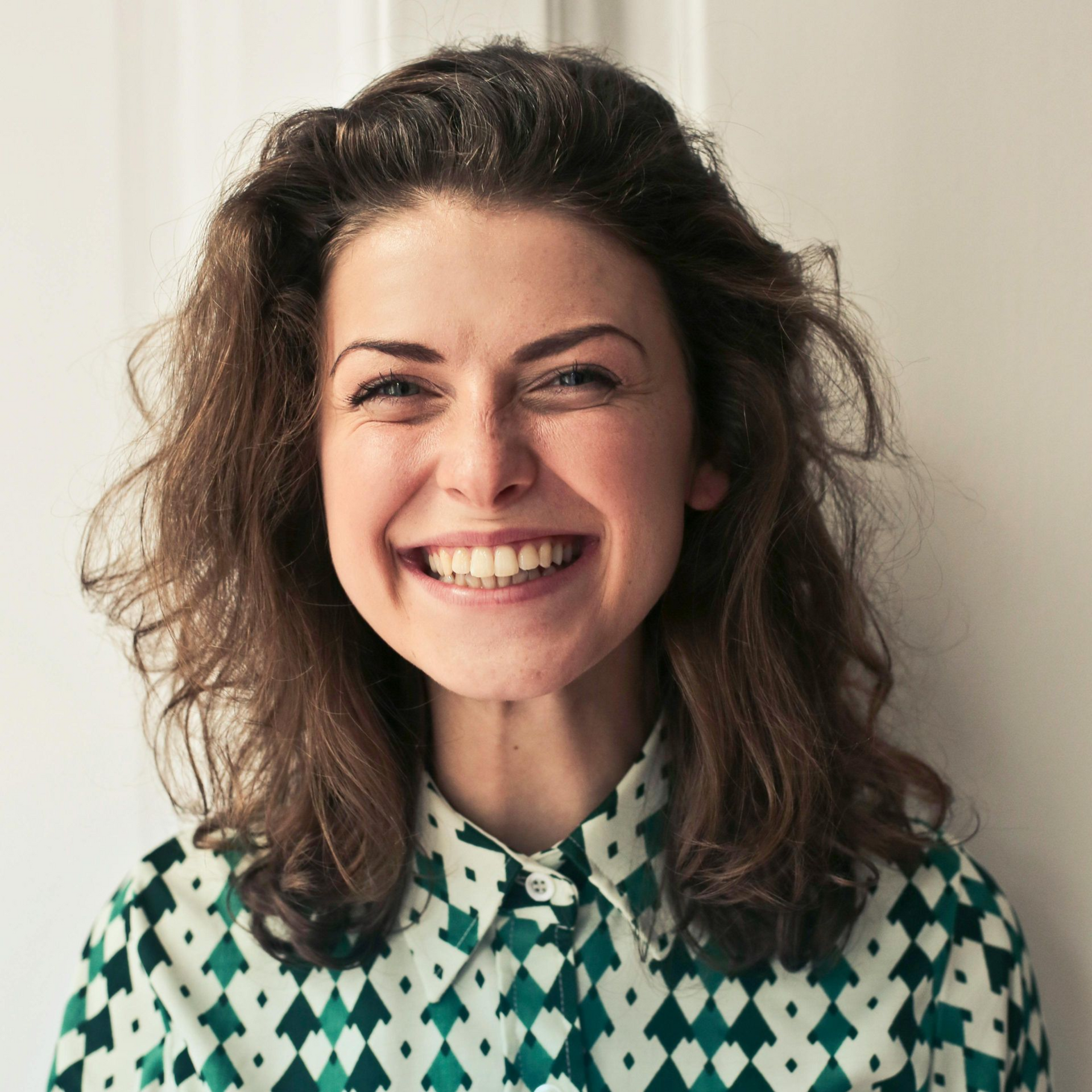 Woman with brown wavy hair smiles broadly, wearing a green geometric patterned shirt, indoors.