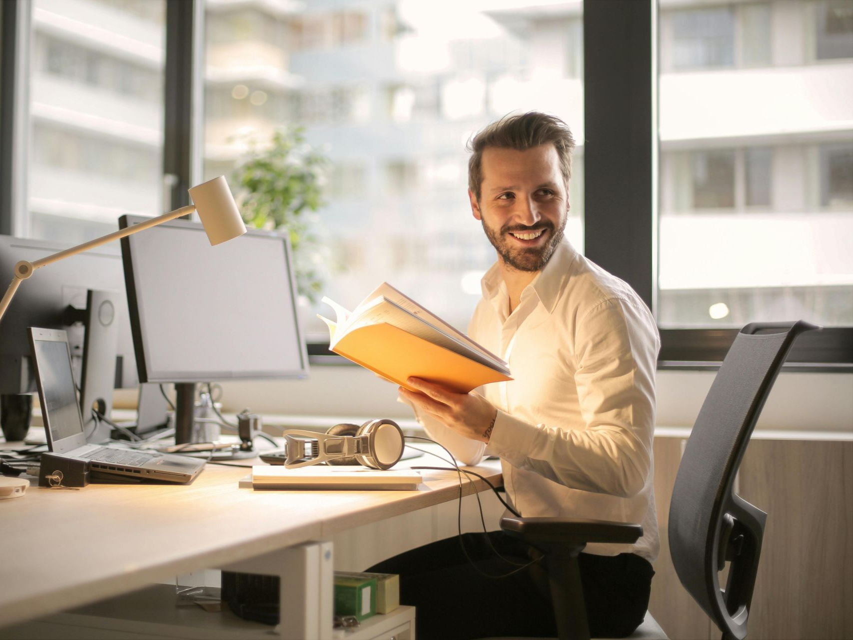 Man at desk smiles, holding a book, with computer and office setting in the background.