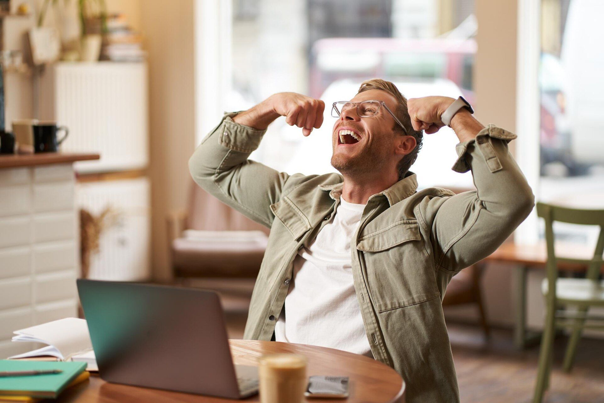 Man at laptop, celebrating success with raised arms and joyful expression, indoors at a cafe.