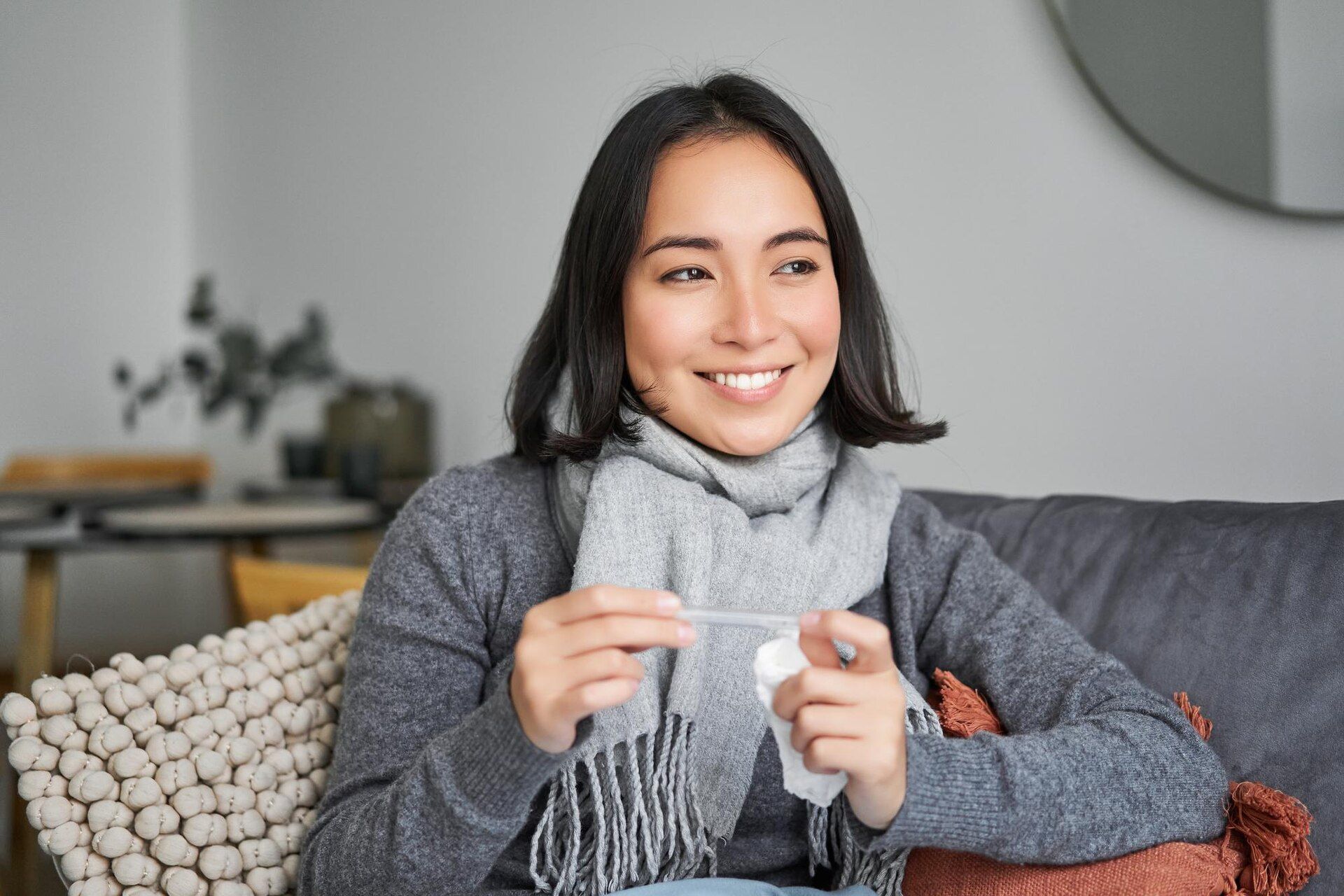 Woman on sofa with thermometer and tissue, wearing scarf, smiling.