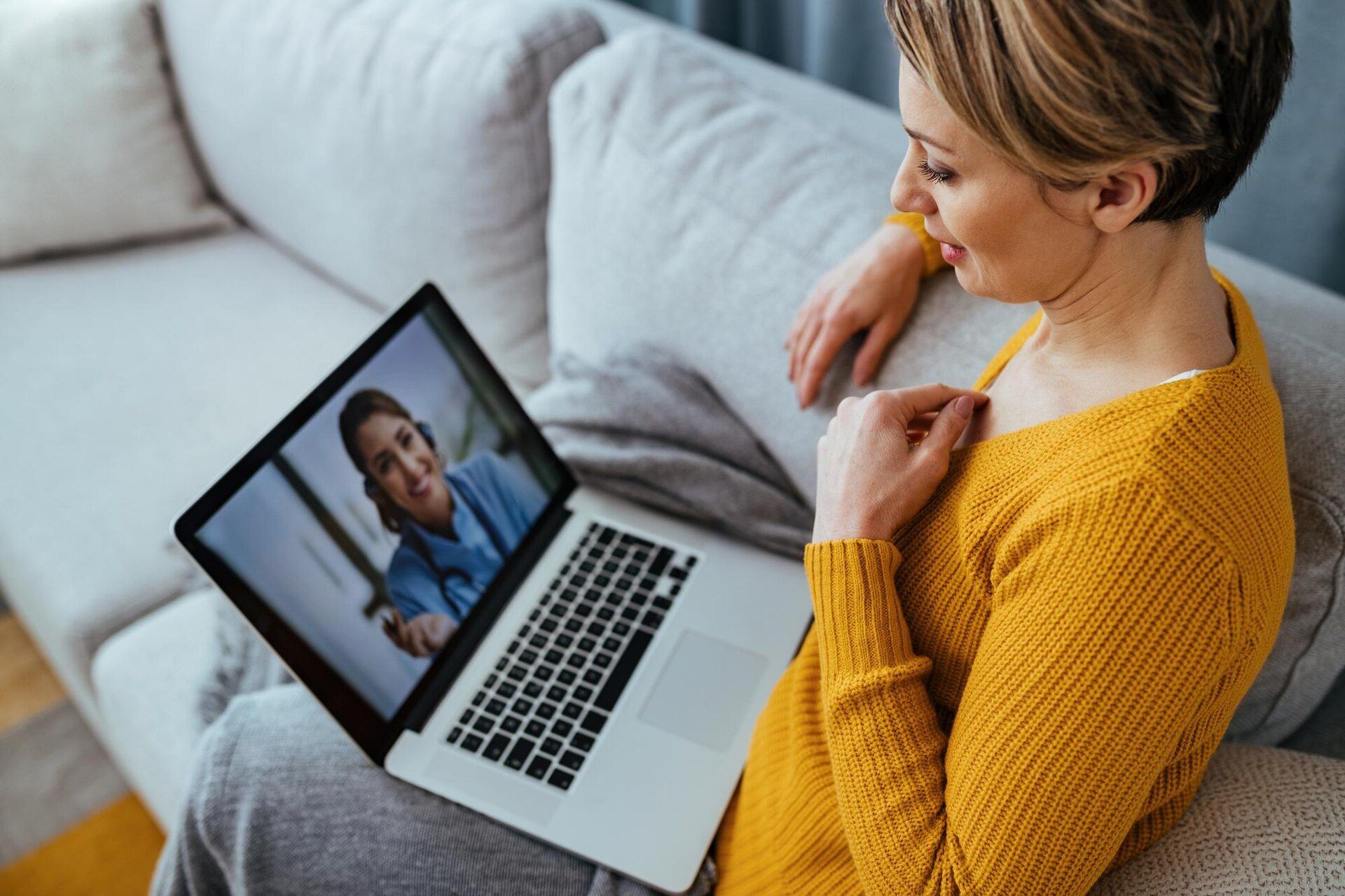 Woman in yellow top on a laptop video call, smiling, seated on a couch.