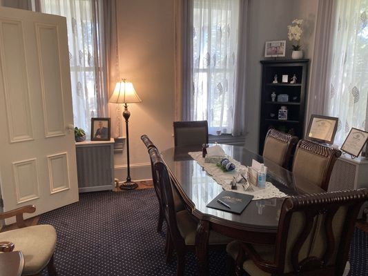 Formal dining room with long table, chairs, lamp, windows, and a bookcase.