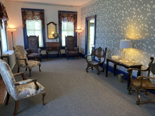 A room with antique furniture, floral wallpaper, two windows with curtains, and two lamps.