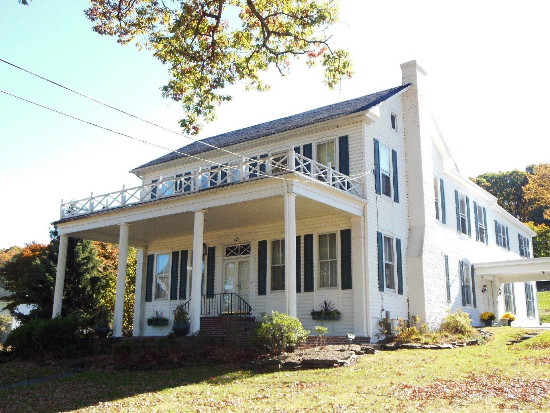 White two-story house with a porch and balcony, green shutters, set on a grassy hill.