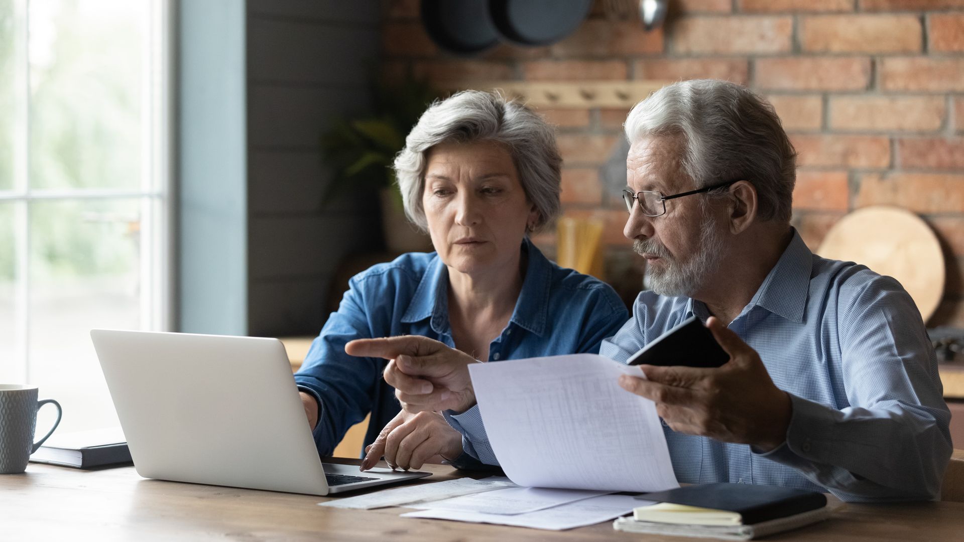 Older couple looking at laptop and paperwork at a table, discussing finances in a kitchen.