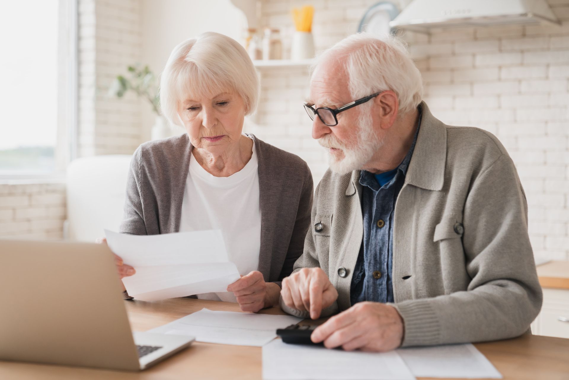 Older couple reviews papers and uses a calculator at a table with a laptop in a well-lit kitchen.