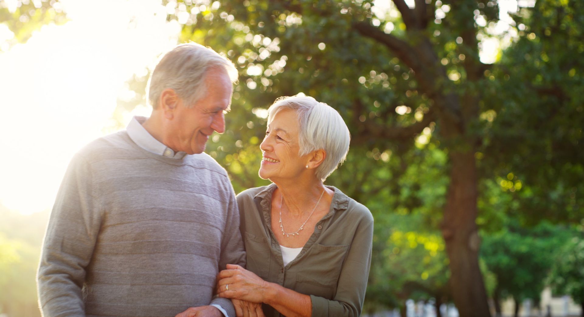 Smiling older couple walking in a park, arm in arm, sunlight in the background.