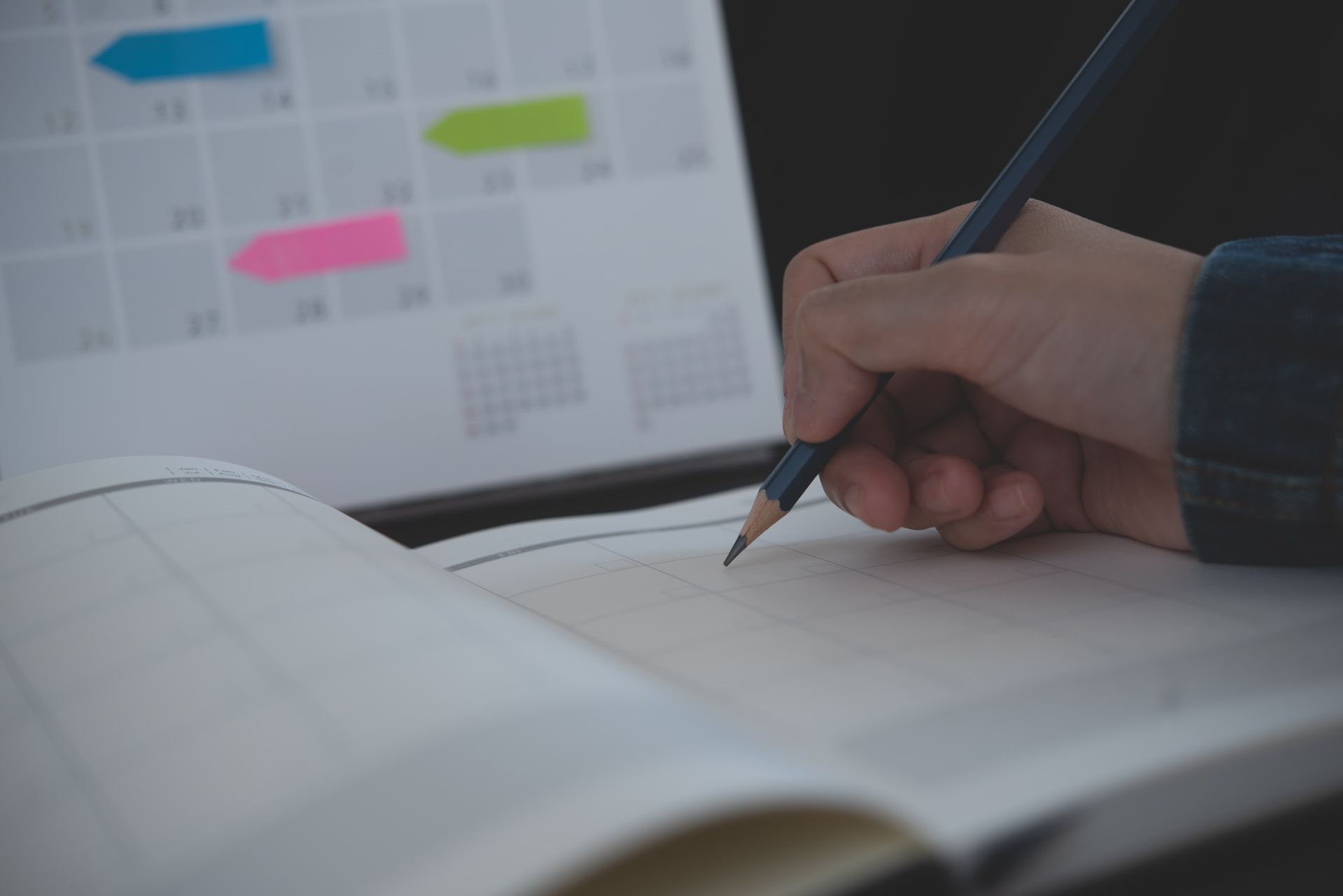 Hand writing in a notebook with a pencil, calendar in the background.
