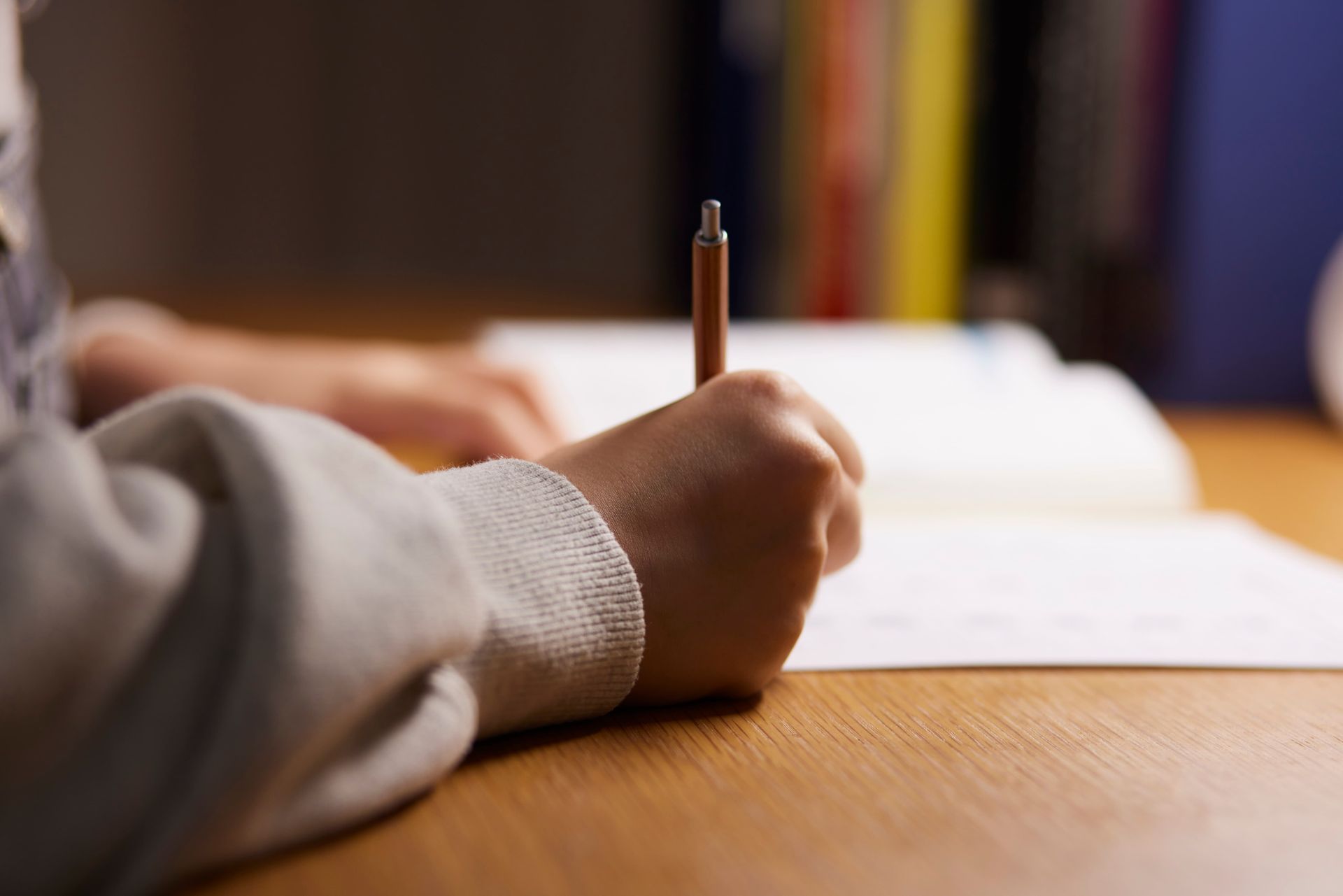 Hand holding a pencil, writing on a paper at a wooden desk, with a blurred book in the background.
