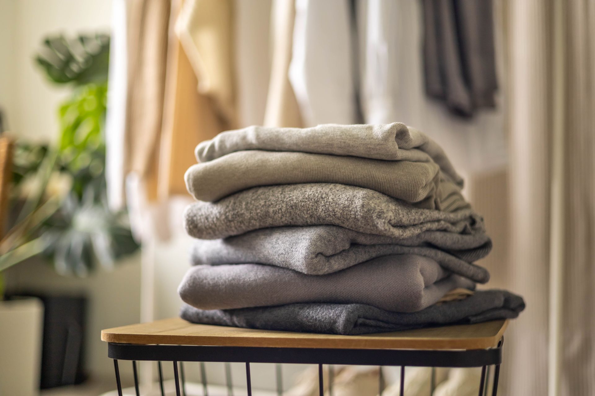 Stack of folded sweaters in shades of gray on a small wooden table, closet background.