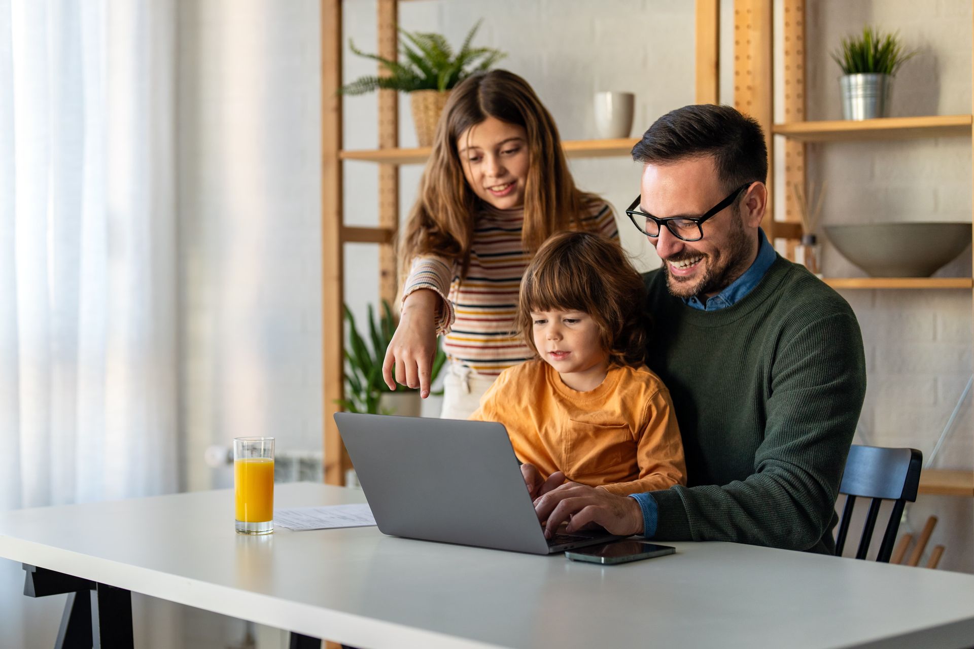Man with glasses and two children looking at a laptop at a white desk in a room with shelves.