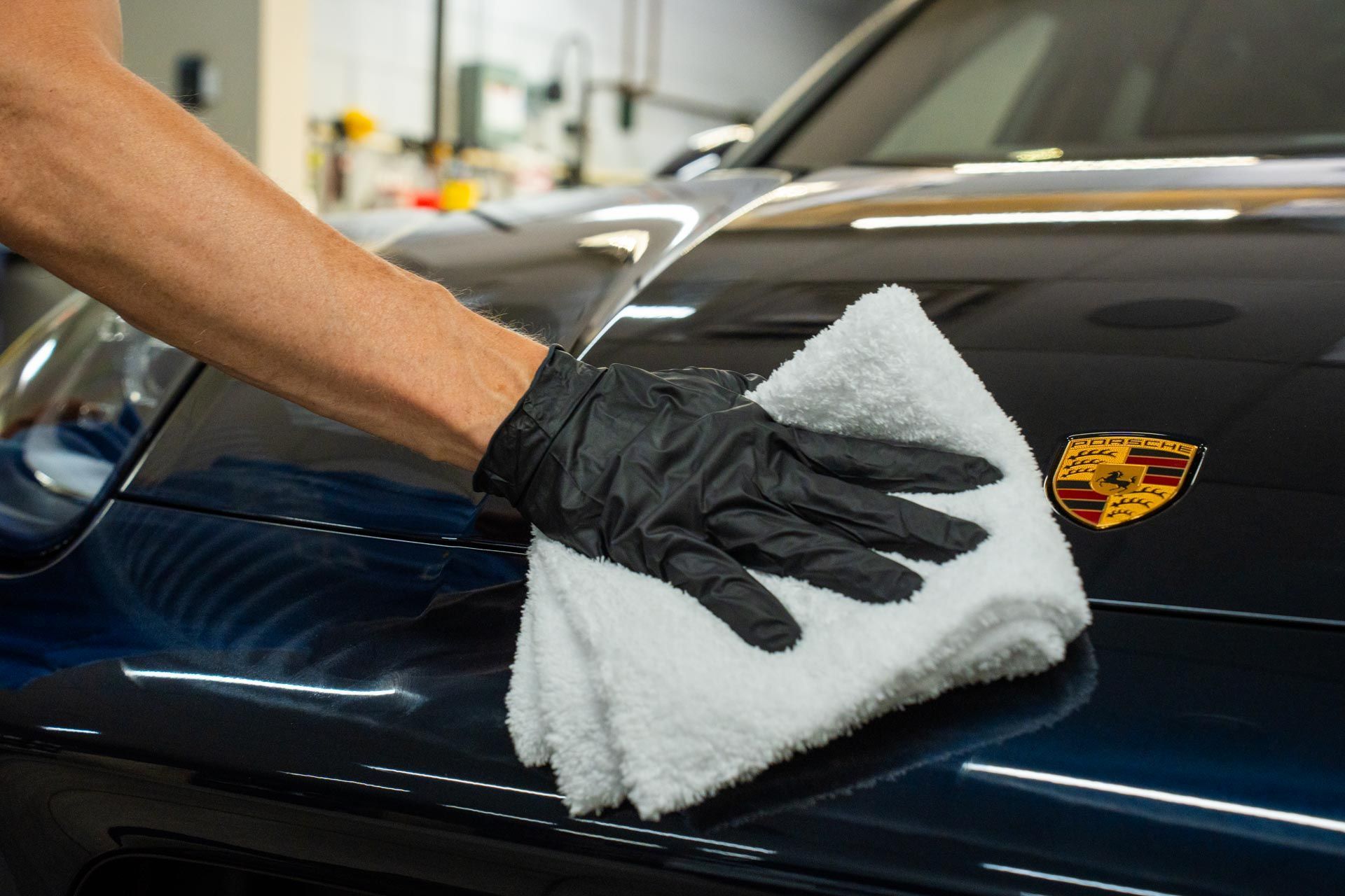 Person in black glove polishing a dark blue Porsche hood with white cloth.