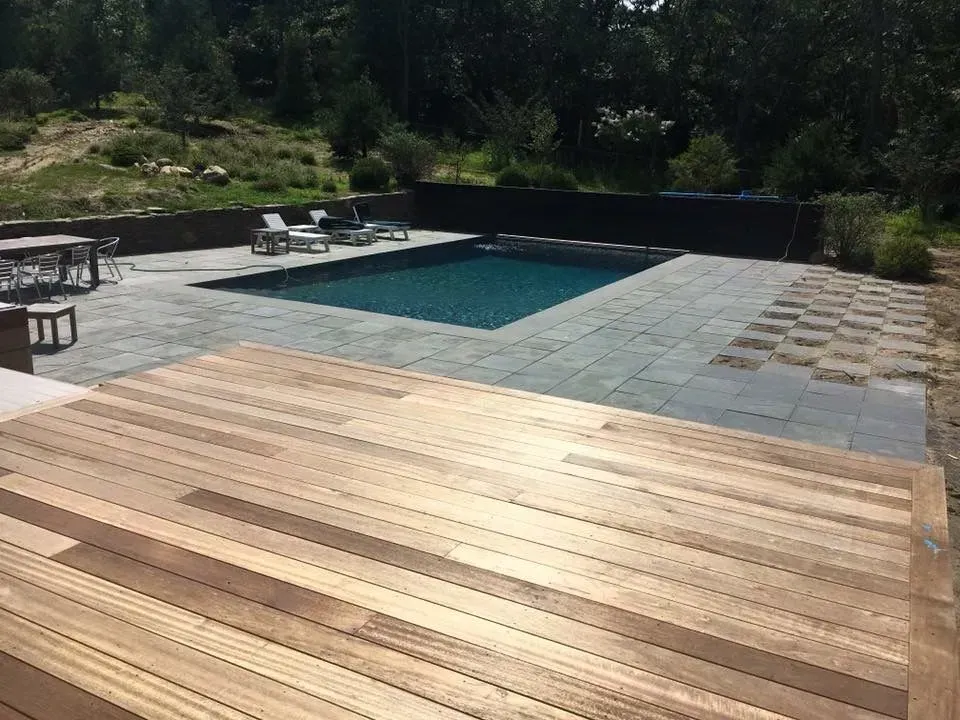 Wooden deck overlooks a pool with lounge chairs, surrounded by stone patio, trees, and foliage.