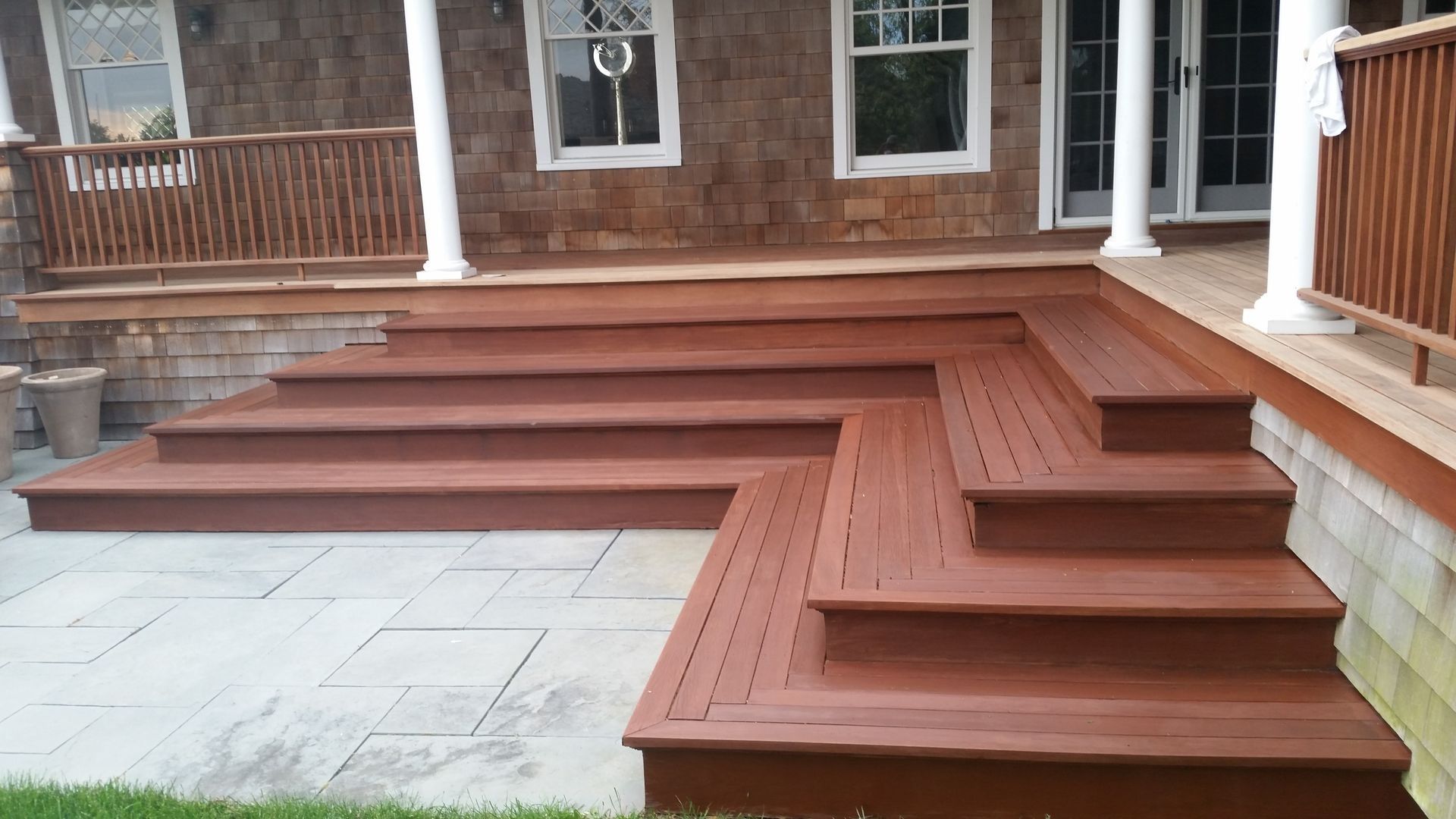 Wooden deck with steps leading down to a stone patio, beside a house with columns and railings.