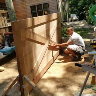 Man building a large wooden door outdoors. He is kneeling, wearing shorts and a white shirt. Sunny setting.