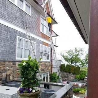 Person on a tall ladder, working on a house with gray siding and a stone base.