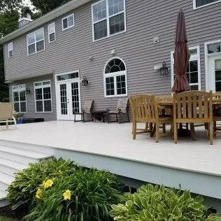 Backyard with a gray house, large white deck, wooden furniture, and green shrubbery.