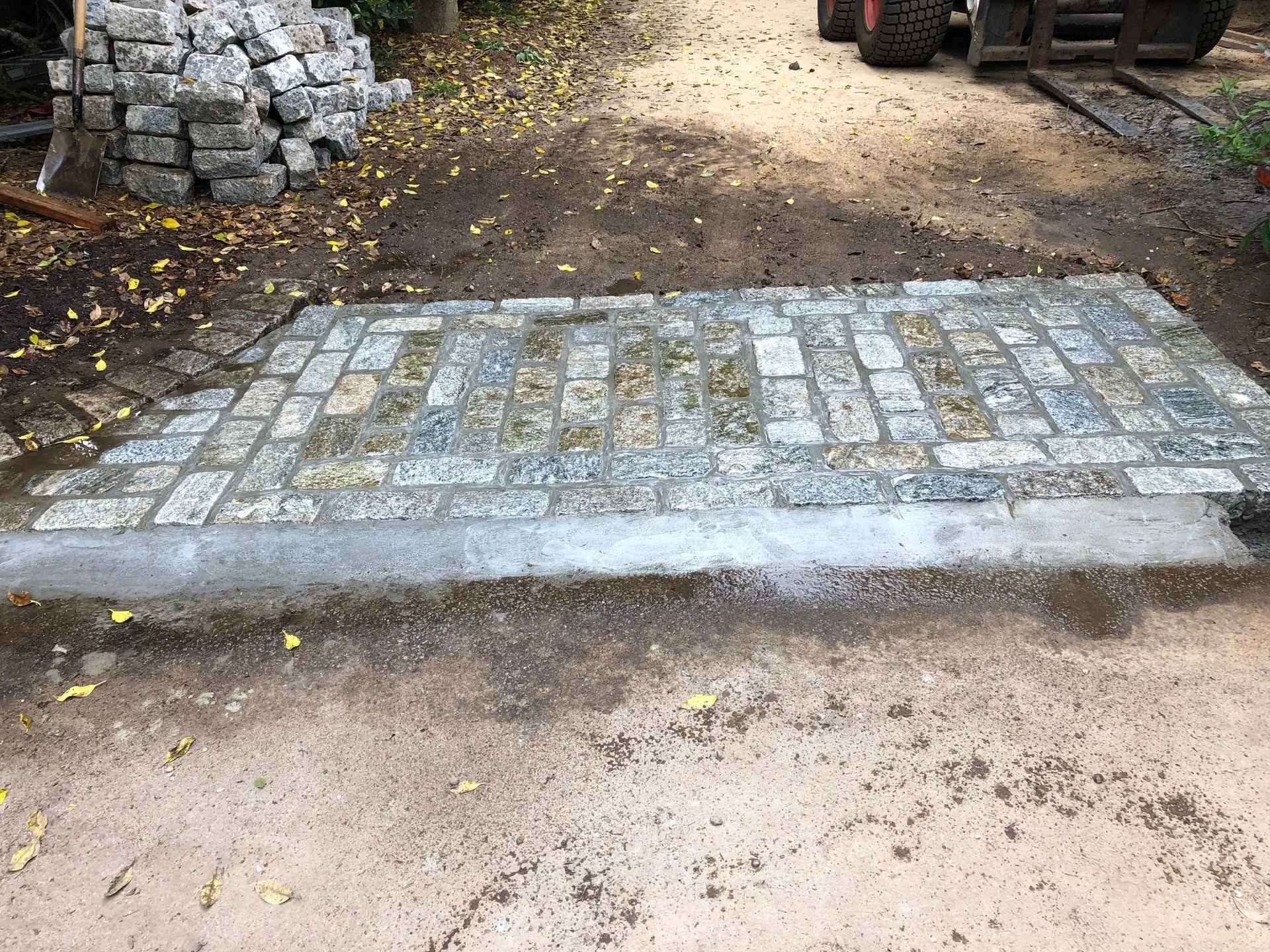 Stone pavers laid over a concrete foundation on a dirt surface with a stack of stones in the background.