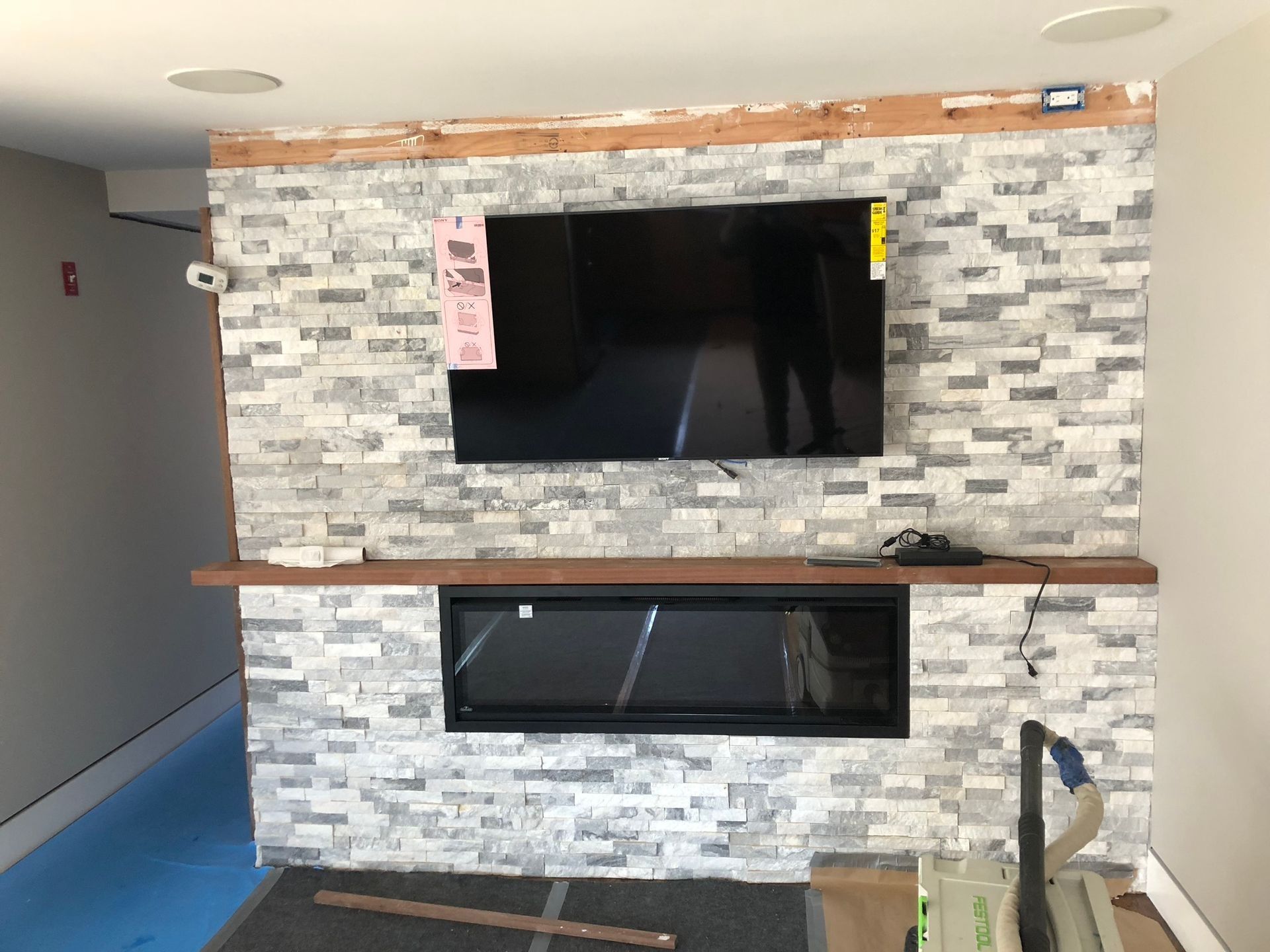 Television and fireplace mounted on a stone wall, with a wooden shelf and exposed ceiling beam.