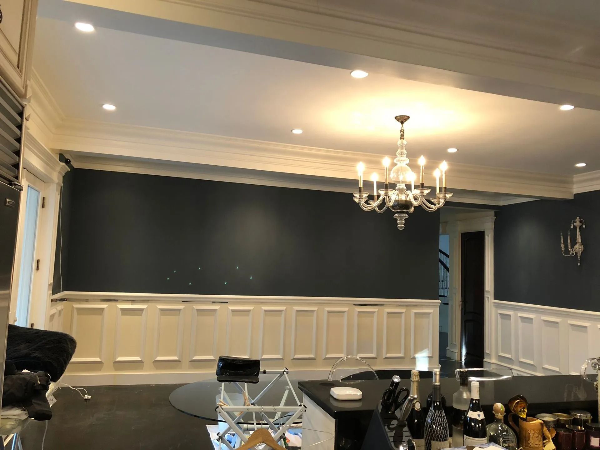 Dining room with blue walls, white wainscoting and ceiling trim. Chandelier hangs over dark countertop.