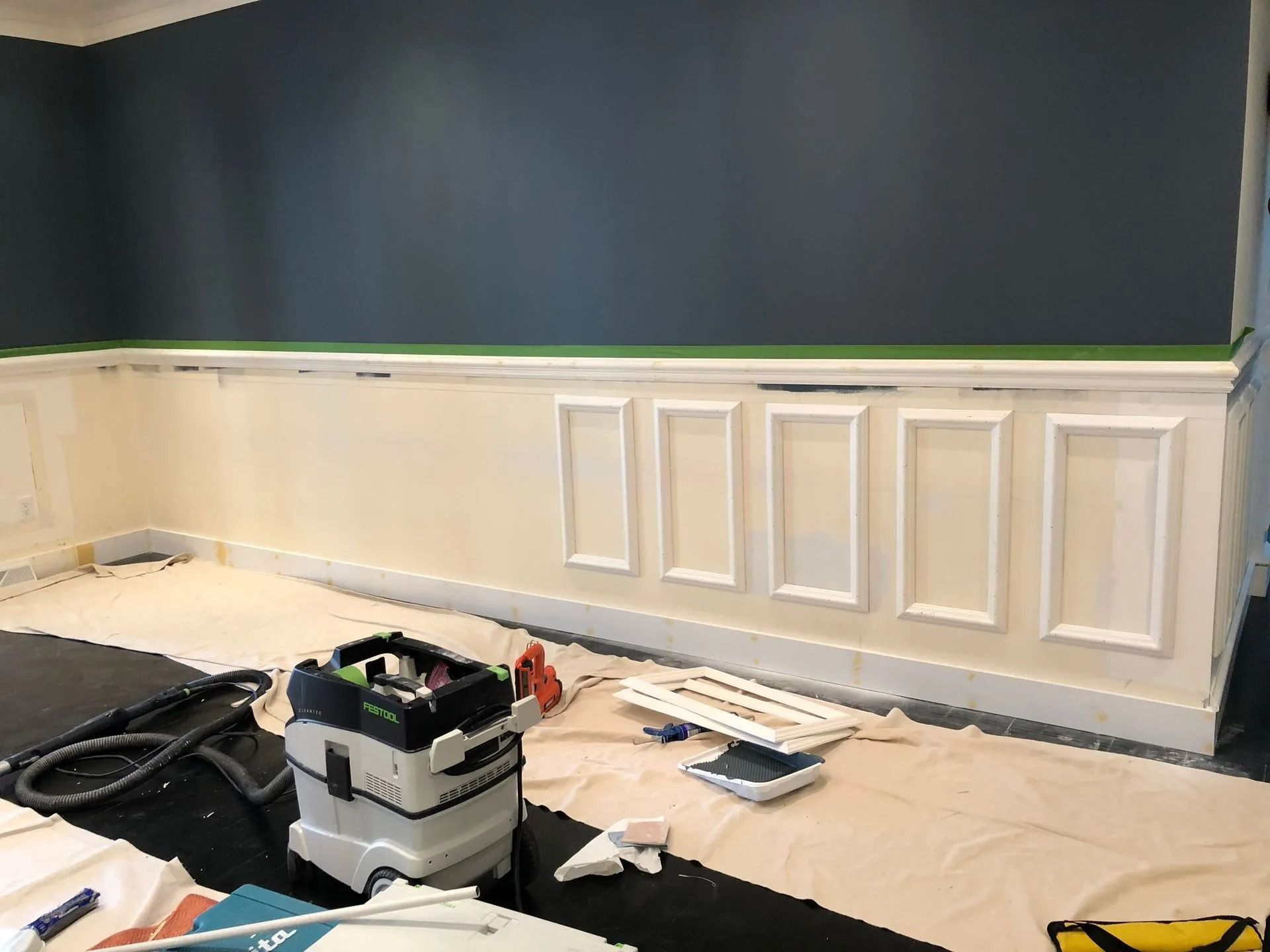 A room with wainscoting being painted. White trim against a dark blue wall. Painting supplies on floor.