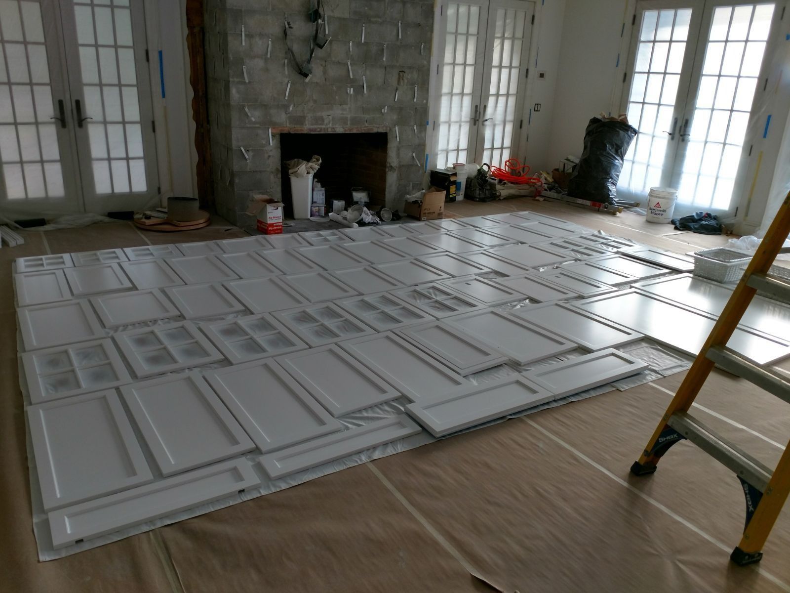 White cabinet doors laid out on a floor covered in protective paper, near a fireplace and ladder.