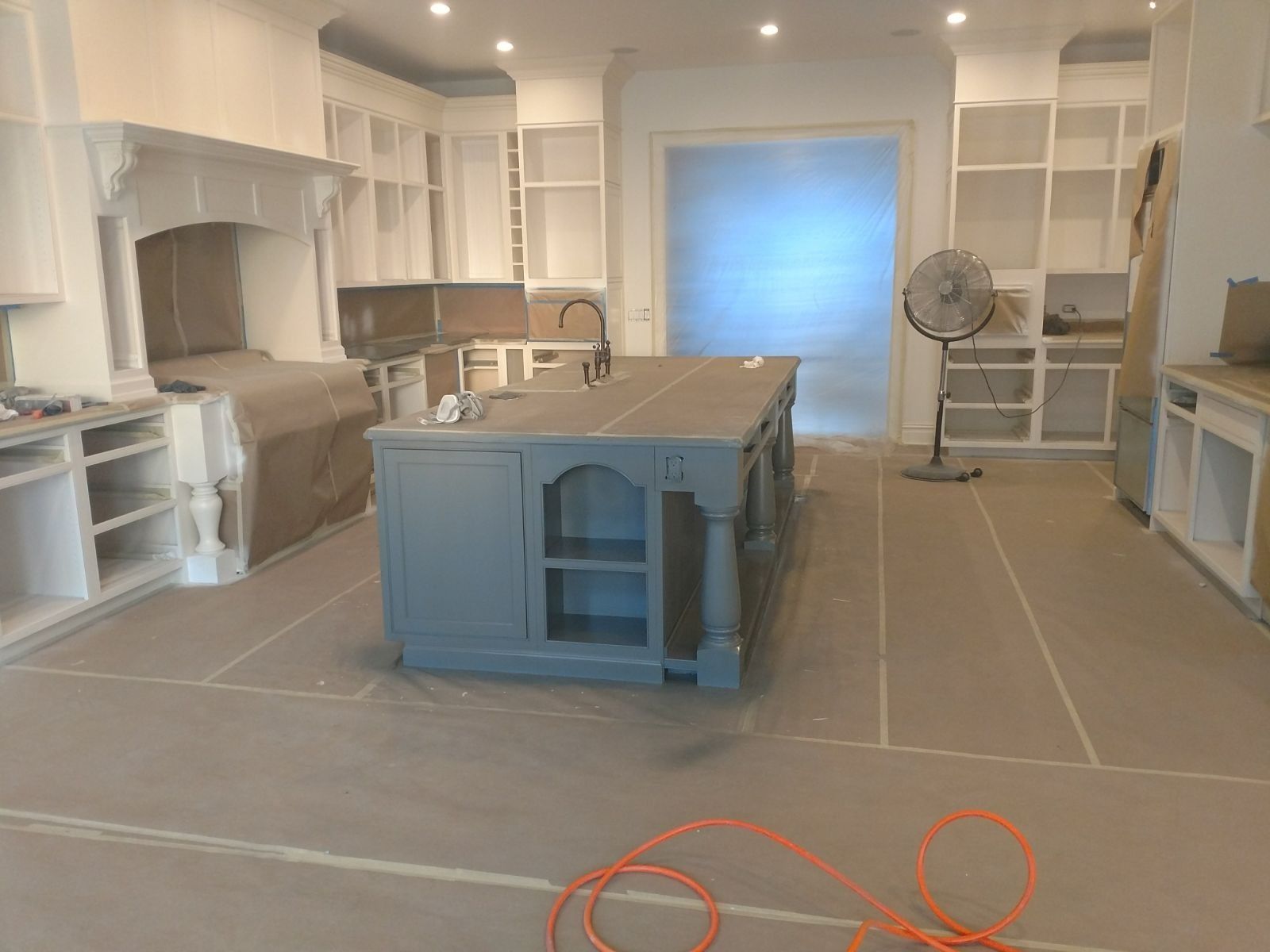 Kitchen undergoing renovation, cabinets painted white and island gray, covered floor, fan running.