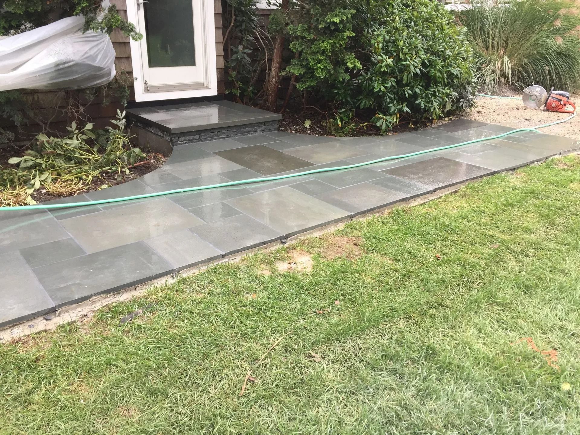Gray stone pathway leading to a doorway, next to a green lawn. A green hose lies on the pathway.