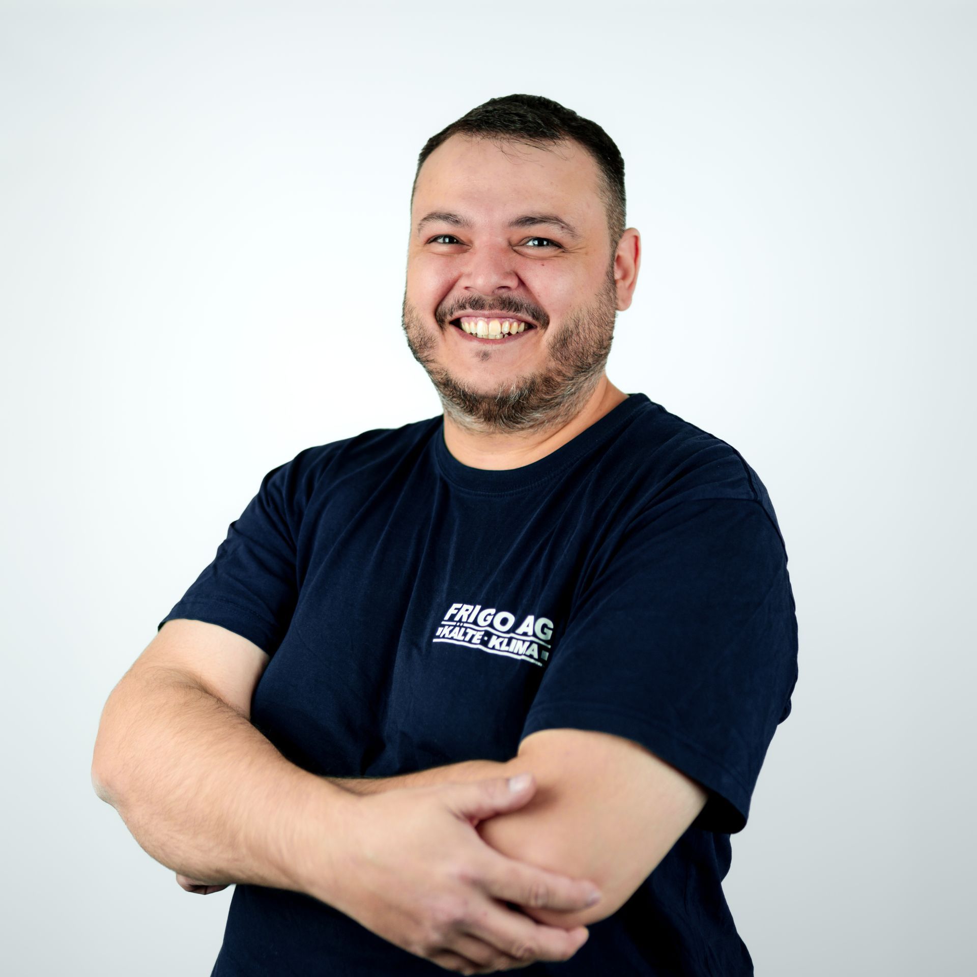 Mann mit verschränkten Armen, lächelnd, trägt ein dunkelblaues T-Shirt mit Logo vor weißem Hintergrund.